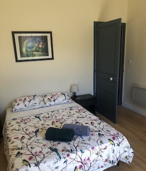 A bedroom with a floral-patterned bedspread, a framed picture on the wall, a bedside lamp, and a dark door. - 3-bedroom apartment in the heart of a medieval village in Vallespir (Pyrenees). (Arles sur Tech)
