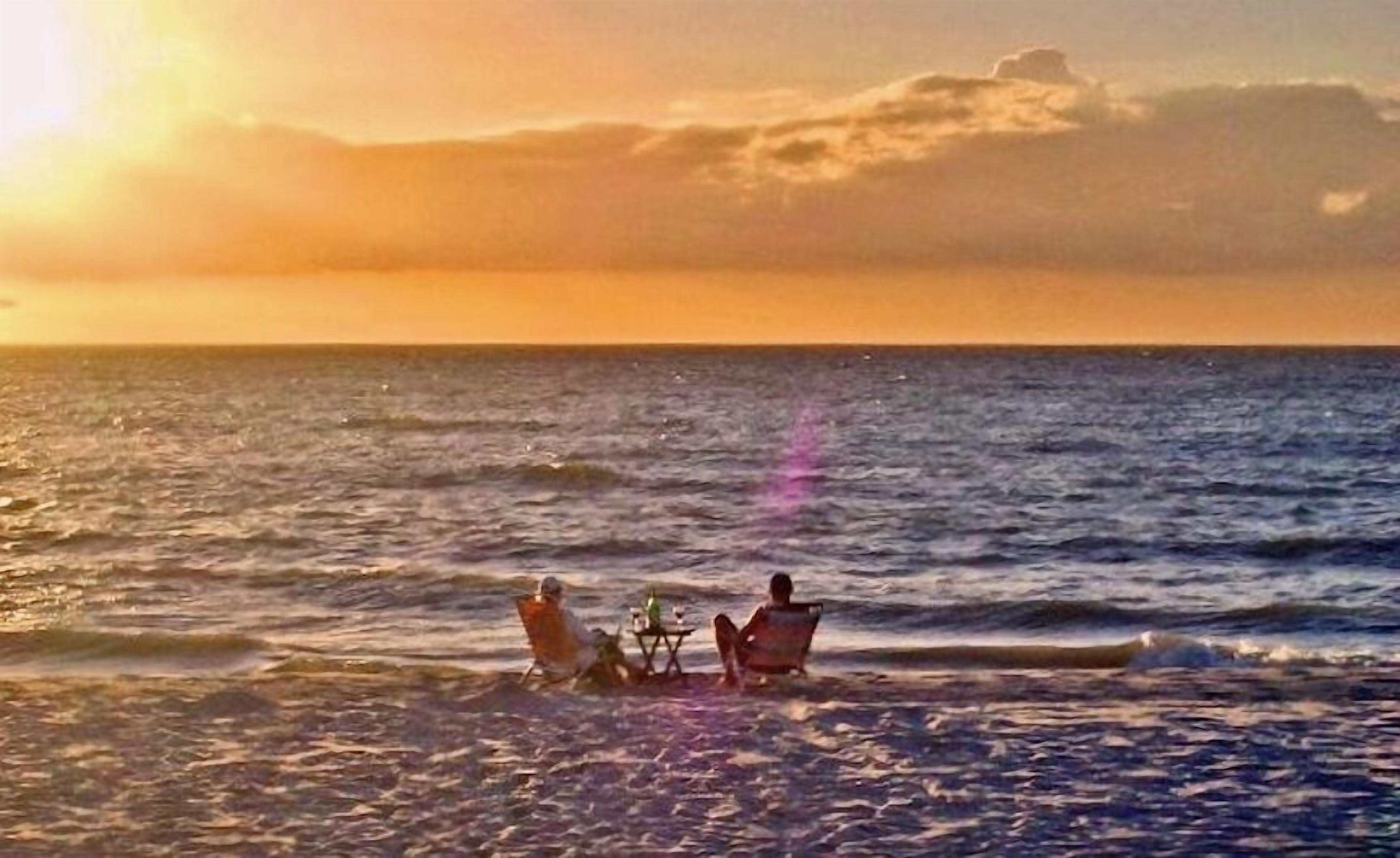 A sunset over the ocean with a person sitting on a chair and another person standing by a table.