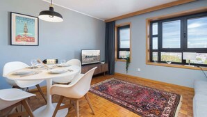 Un espace repas moderne avec une table ronde, des chaises blanches, un luminaire suspendu noir et un tableau encadré représentant le Portugal.