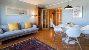 Un salon avec un canapé gris, des coussins jaunes, un tapis à motifs, une table à manger avec des chaises blanches et un tableau encadré au mur.