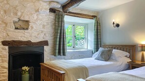 A bedroom with a stone wall, a fireplace, two beds, a window with curtains, and a nightstand with a lamp. - 2 bedroom accommodation in Satterthwaite, near Coniston (Satterthwaite, near Coniston)