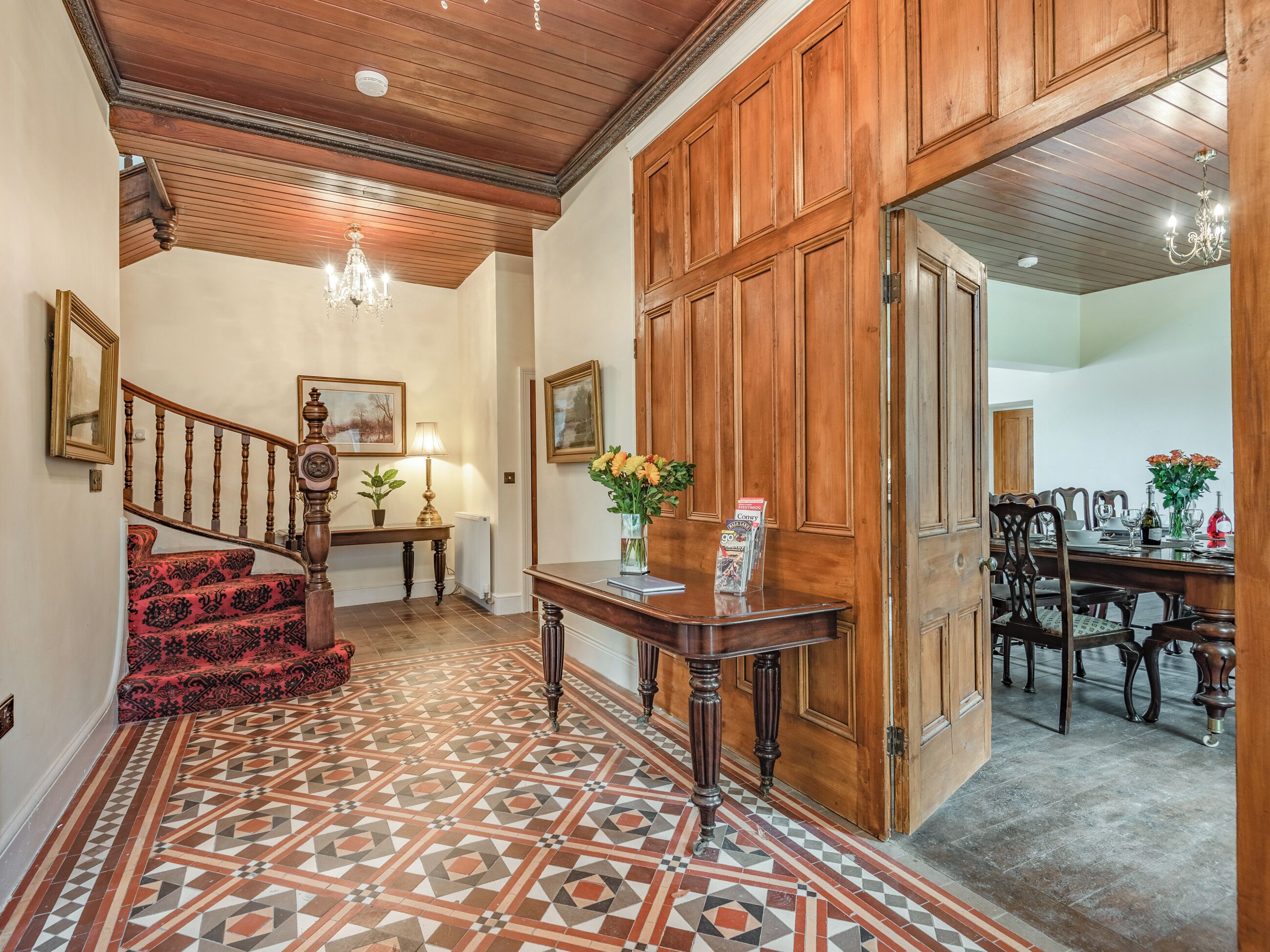 Un hall espacioso con piso de baldosas estampadas, una escalera de madera, una mesa de recepción y un comedor con muebles de madera.