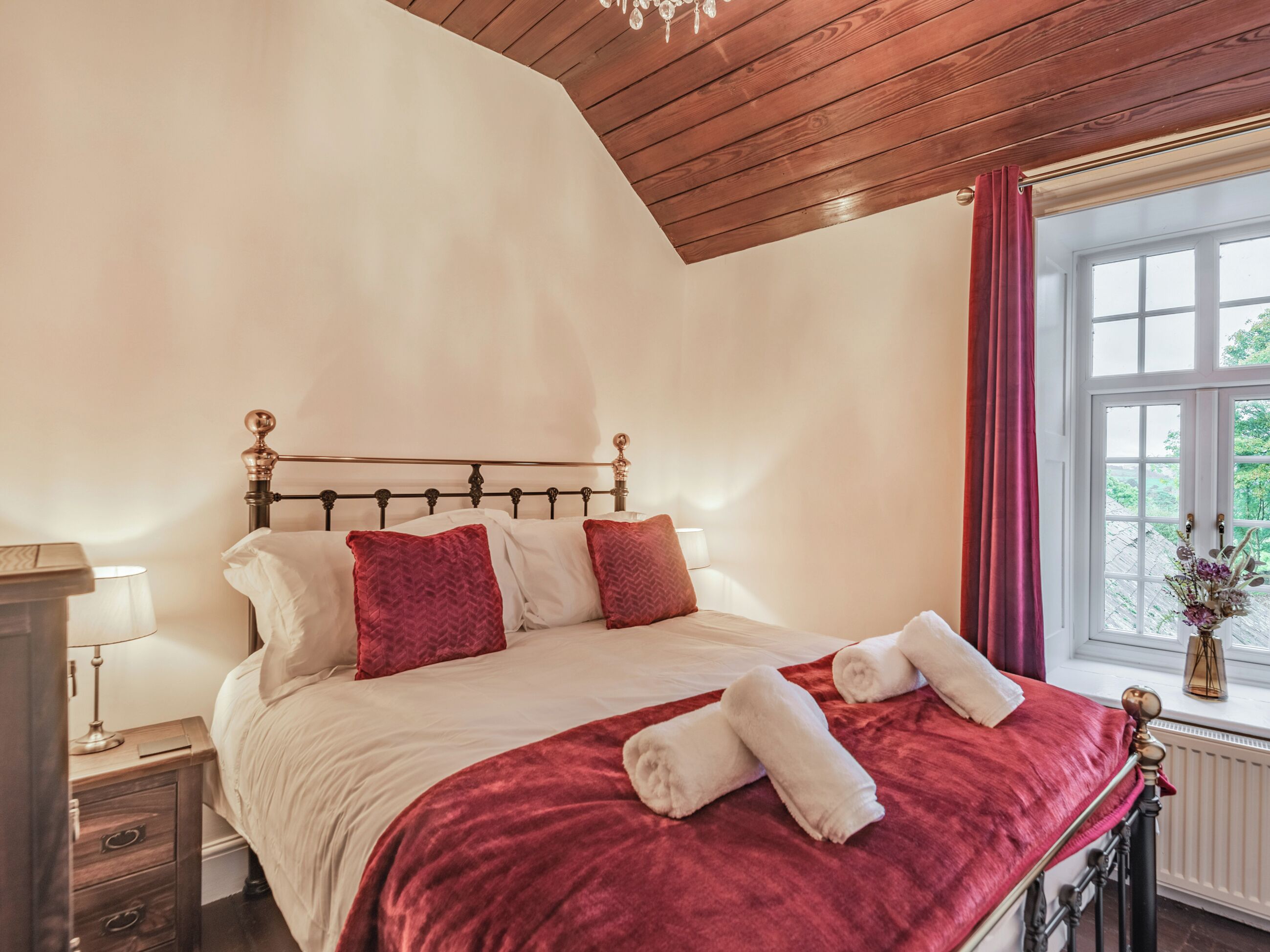Une chambre avec un plafond en bois, un lit recouvert de draps blancs et agrémenté d’oreillers rouges, un plaid rouge et une fenêtre donnant sur un paysage verdoyant.