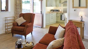 A living room with a brown armchair, a red sofa, and a coffee table with pastries. There is a kitchen in the background with light green cabinets and a microwave. - 1 bedroom accommodation in Newton Le Willows (Newton Le Willows)