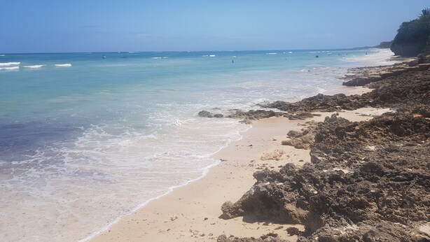 Uma praia com águas turquesa cristalinas, areal arenoso e afloramentos rochosos.