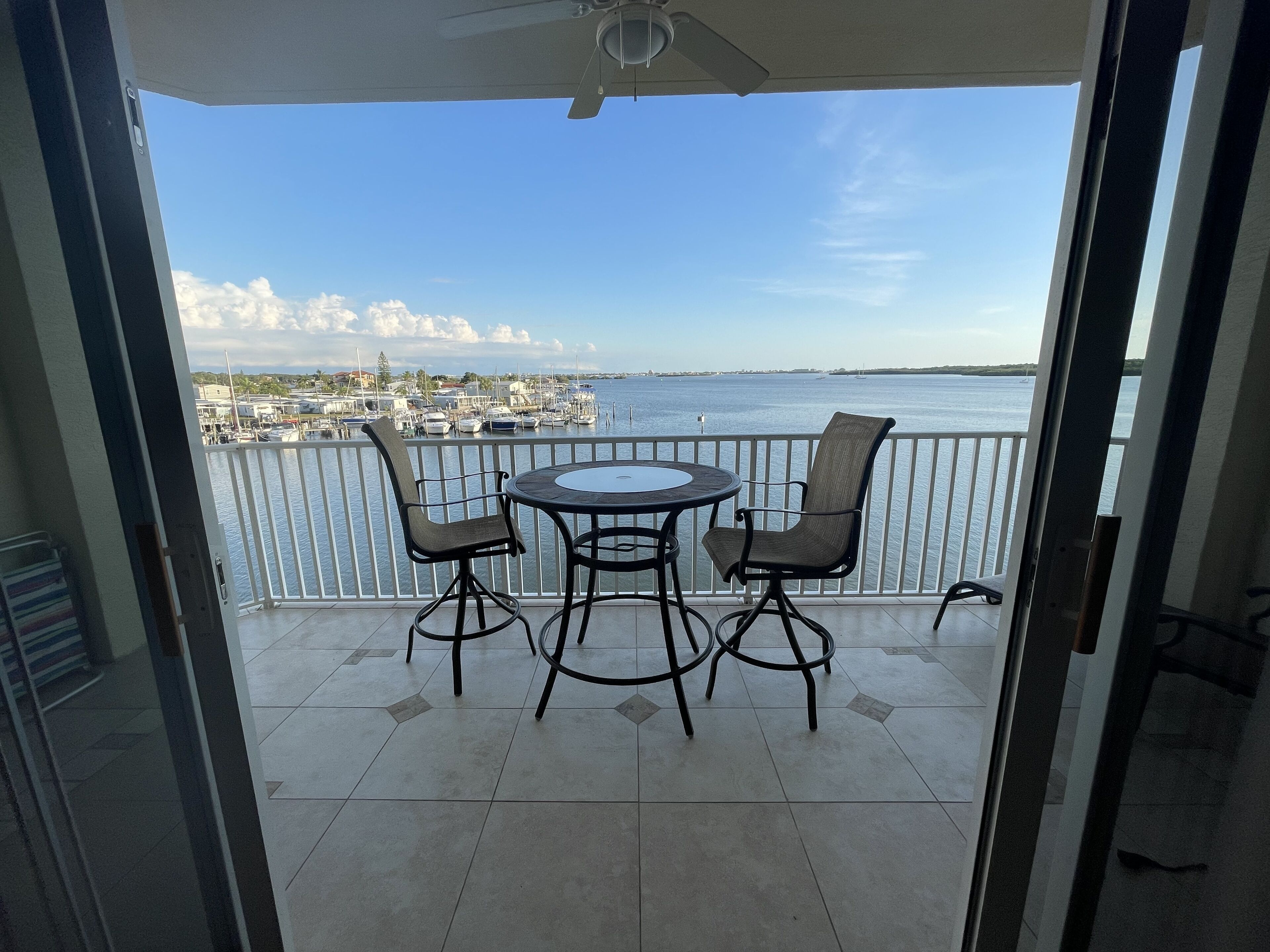 A balcony with a table and two chairs, overlooking a marina with boats and a distant cityscape.