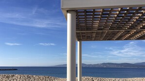 A beachside seating area with a pergola, benches, and chairs, overlooking a calm sea and distant hills.
