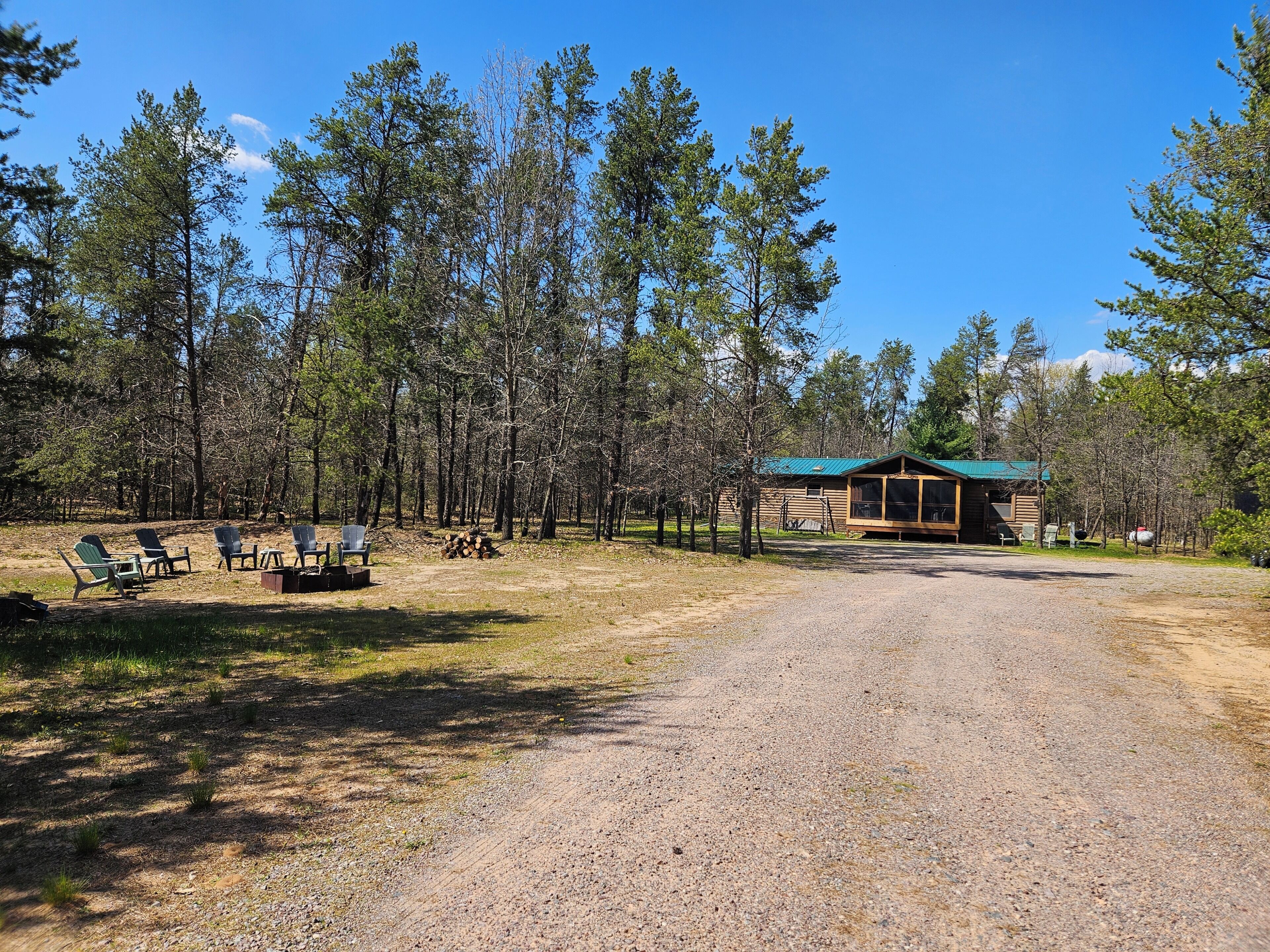 A rustic cabin with a gravel driveway, surrounded by tall trees and a clear sky.