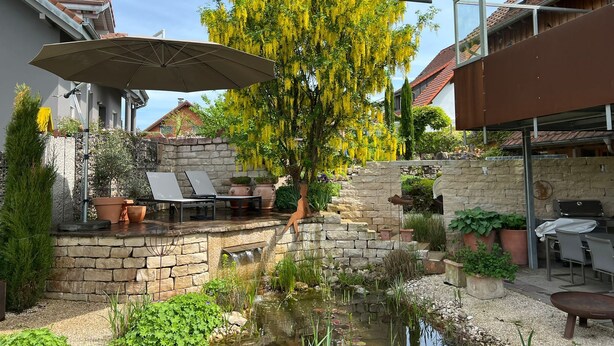 A patio area with a stone wall, a small pond, and a tree with yellow flowers. There are outdoor furniture pieces and a grill. - Small black forest lodge (Efringen-Kirchen)