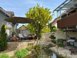 Een terras met een stenen muur, een kleine vijver en een boom met gele bloemen. Er staan tuinmeubels en een barbecue.