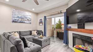 A modern living room with a grey sectional sofa, a small table with a colorful chair, a fireplace, and a large TV. - Desert retreat, 3 pools, free parking, PAC MAN, Pickleball, Basketball, Internet (Phoenix)