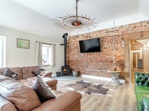 A living room with a brick wall, a fireplace, a leather sofa, a green chair, and a mounted TV. - Cattle Crush Cottage - UK35988 (Skerne, near Driffield)