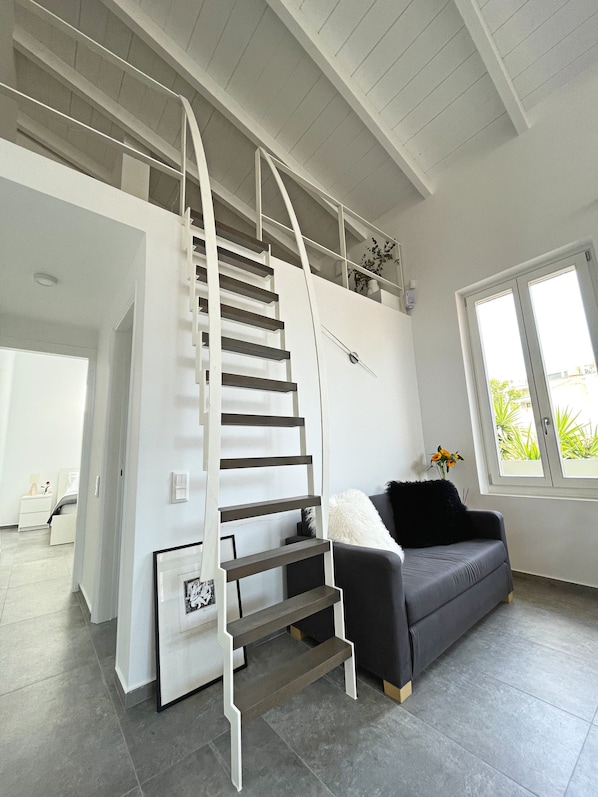 Ein modernes Interieur mit einer Treppe zu einer oberen Ebene, einem grauen Sofa und einem großen Fenster mit Blick auf Grünflächen.