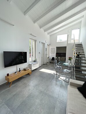 A spacious living area with a large flat-screen TV, a wooden console table, and a dining area with clear chairs. - Unique Flat (Athens)
