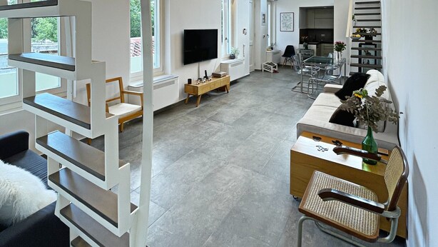 A spacious living area with a high ceiling, built-in shelving, a flat-screen TV, and a staircase leading to an upper level. - Unique Flat (Athens)