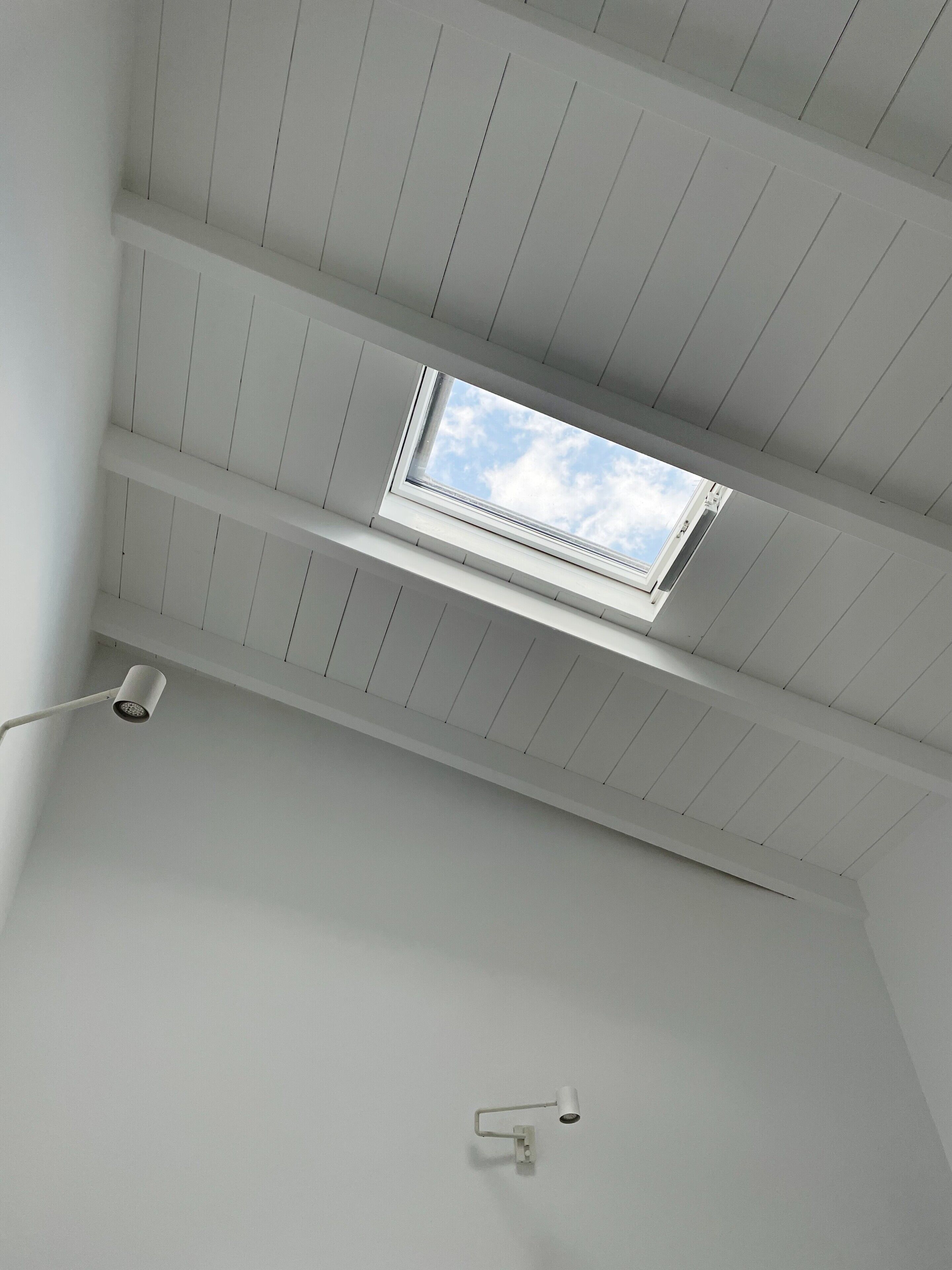Une lucarne dans une pièce aux murs blancs et au plafond orné de panneaux de bois disposés horizontalement.