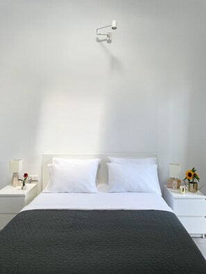 A modern bedroom with a large bed, white nightstands, a wall-mounted lamp, and a vase with flowers. - Unique Flat (Athens)