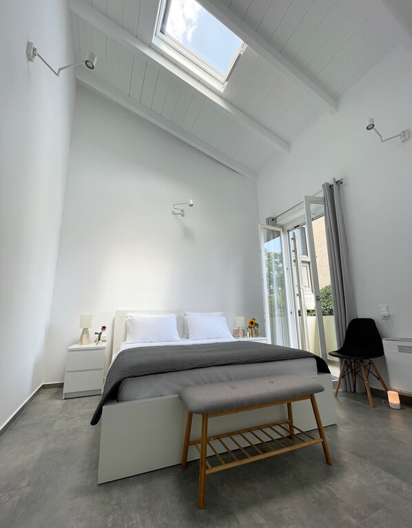 A modern bedroom with a skylight, a bed with a grey blanket, a bedside table with a lamp, and a bench. - Unique Flat (Athens)