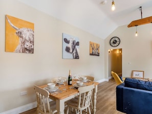 A dining area with a wooden table set for four, surrounded by white chairs. There are paintings on the wall depicting animals. A clock is visible on the wall, and a sofa is in the background.