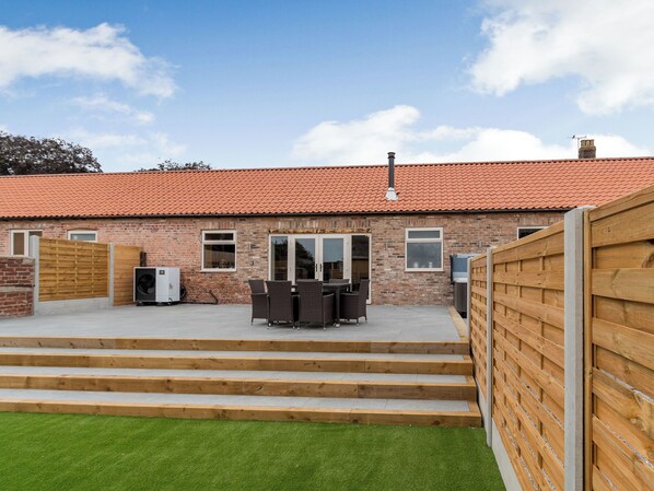 A brick building with a red-tiled roof, a wooden fence, and a patio area with steps leading to a table and chairs.