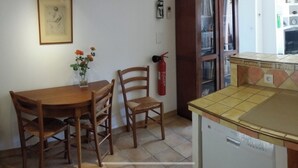 Eine kleine Küche mit einem Holztisch, zwei Stühlen, einer Blumenvase, einem Feuerlöscher und einem Kühlschrank.