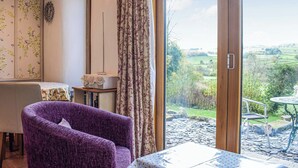 A room with a purple armchair, a table with a floral tablecloth, a small table with a white object, a wall with floral wallpaper, and a large sliding glass door leading to an outdoor garden. - 1 bedroom accommodation in Llansilin, near Oswestry (Llansilin, near Oswestry)