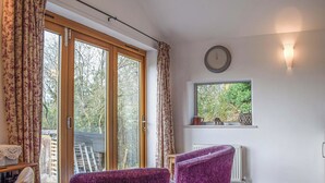 A room with a sliding glass door, a window with a view of greenery, a clock on the wall, and two purple chairs. - 1 bedroom accommodation in Llansilin, near Oswestry (Llansilin, near Oswestry)