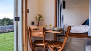 Een kleine kamer met een houten bureau, een stoel, een bed en een tafeltje met wijnglazen en een bloemenvaas.