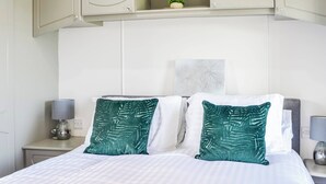 Een net opgemaakt bed met witte beddengoed en twee groene kussens, een nachtkastje met een lamp en een kleine plant erop.