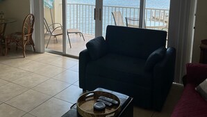 Una sala de estar con un gran ventanal corredero que da a un balcón con vistas al agua. Hay un sofá azul oscuro, una mesa de centro de madera con una bandeja y un cojín rojo.