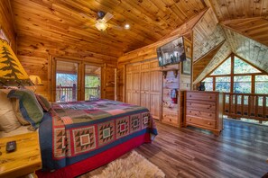 A log cabin bedroom with a large bed, wooden walls, a ceiling fan, and a TV mounted on the wall.