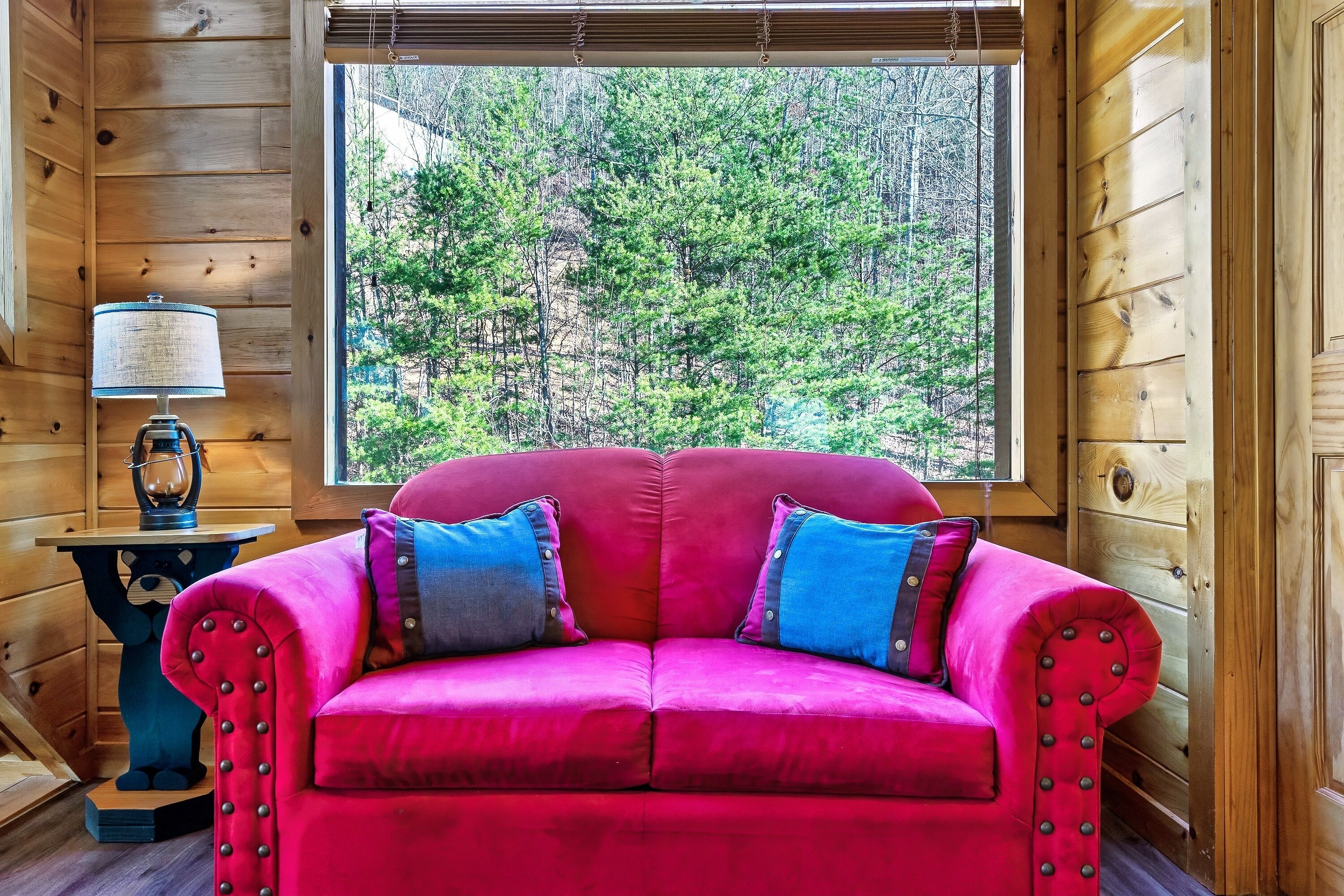 Un canapé en velours rouge agrémenté de deux coussins, une lampe de table et une vue sur une forêt par la fenêtre.