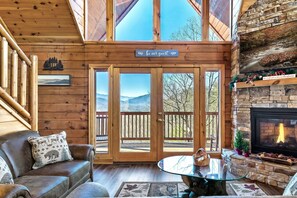 A cozy cabin interior with a fireplace, a wooden staircase, and a scenic view through the doors. - Amazing Views, Secluded, 3 King suites, Sleeps 11, Hot tub. Pet Friendly (Sevierville)