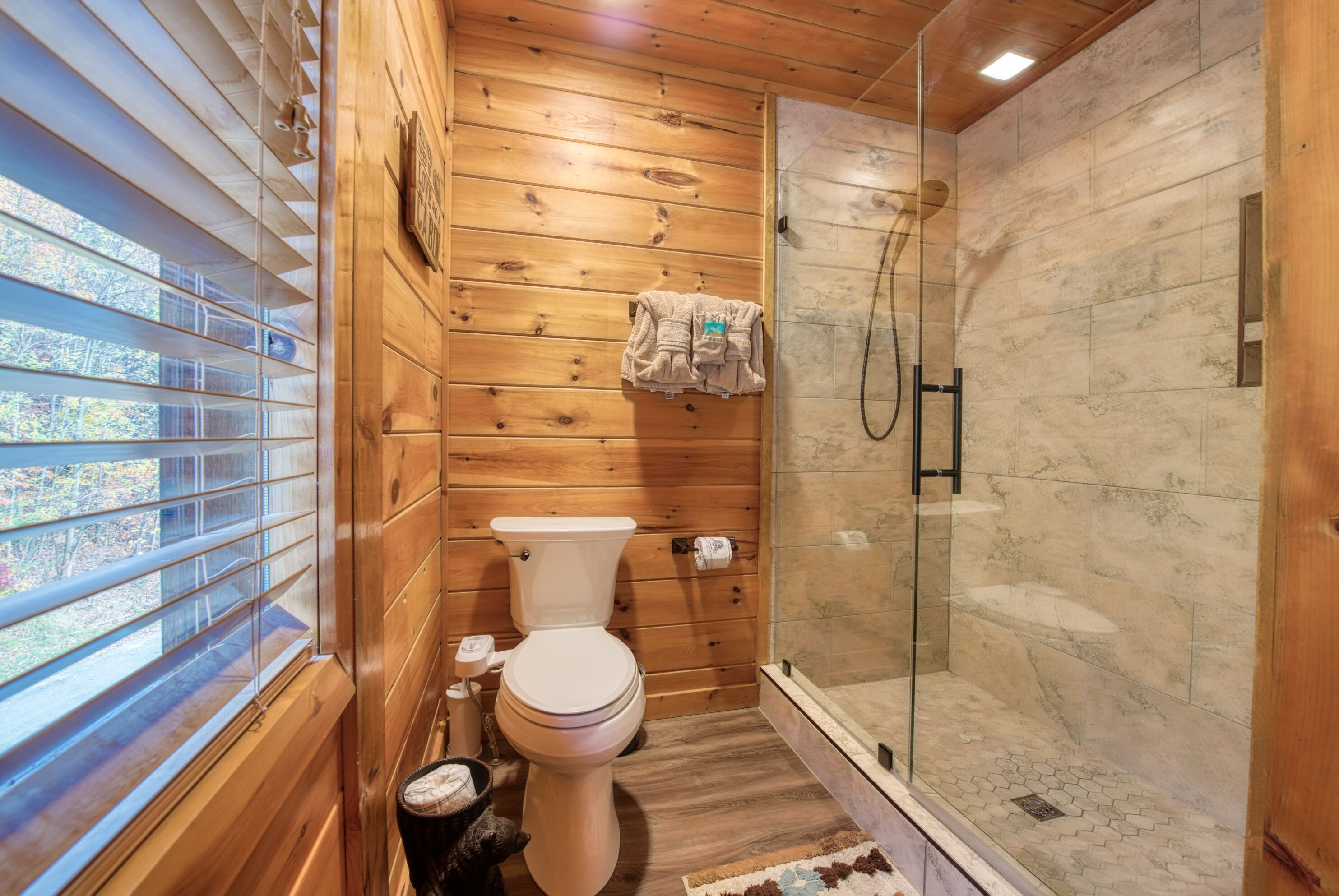 Une salle de bain aux murs lambrissés, équipée de toilettes, d’une douche à parois de verre et d’une fenêtre avec des stores.