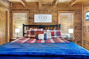 A wooden cabin bedroom with a bed, a sign saying "STAY awhile", and a window with blinds.