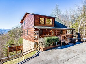A log cabin with a porch, a stone foundation, and a view of the surrounding landscape.