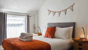 A neatly made bed with a grey headboard, a red throw blanket, and a folded towel on the foot of the bed. There is a bedside table with a lamp and a small plant. - St Martins Square Apartment 3 (Scarborough)