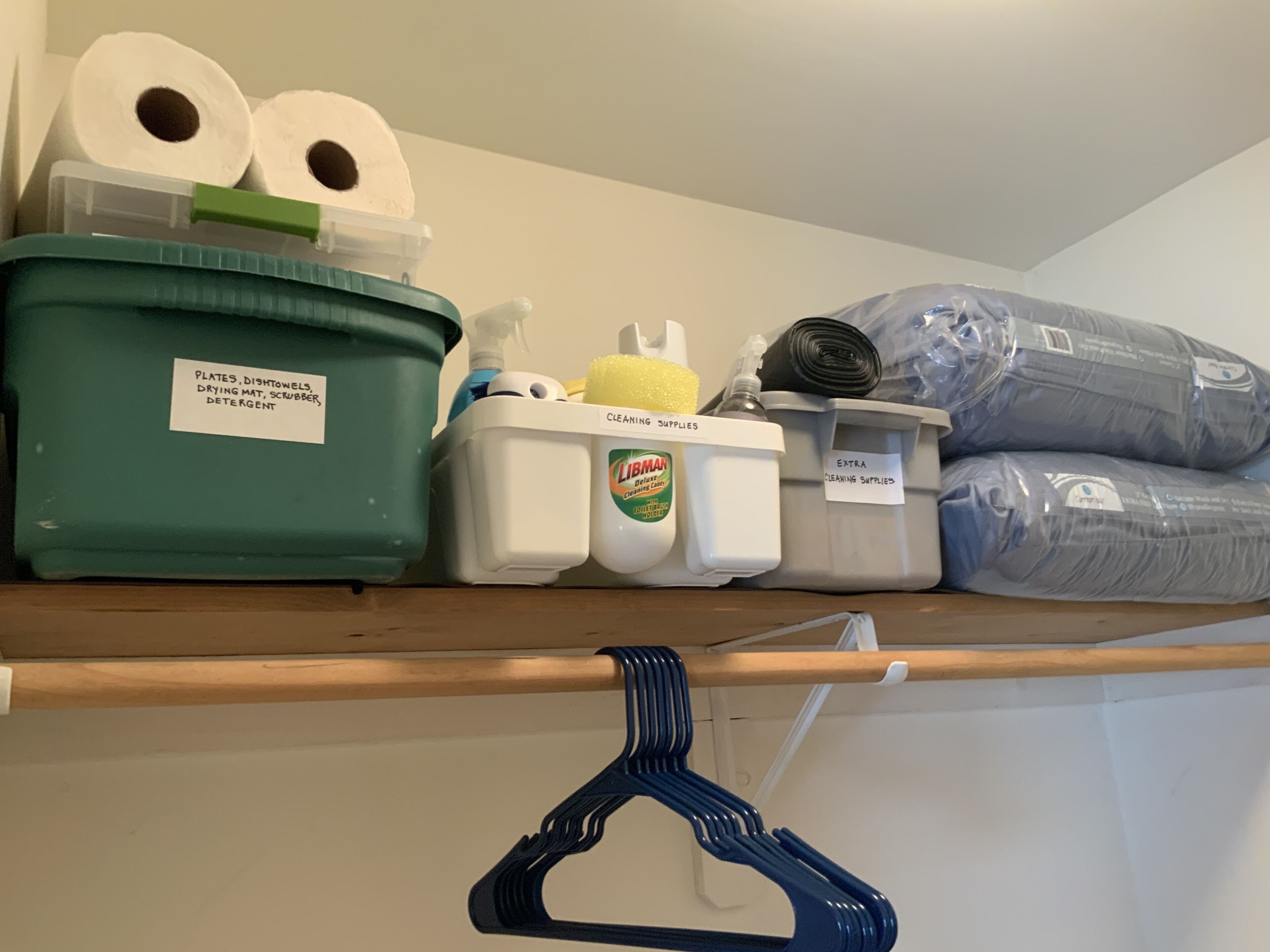 Un área de almacenamiento con cajas etiquetadas que contienen productos de limpieza, un perchero azul colgante y un rollo de papel higiénico.