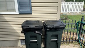 Deux poubelles noires sont placées devant une maison dont le revêtement extérieur est de couleur beige et qui est bordée d’une clôture blanche.