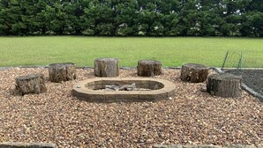 Un foyer circulaire, entouré de pierres et de troncs d’arbres, est aménagé dans un espace recouvert de gravier et bordé de briques.