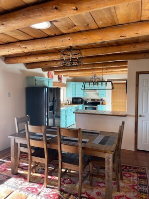 A wooden dining table with chairs, a kitchen with teal cabinets, and a hanging light fixture. - Relaxing Arroyo Seco Vacation Home with Beautiful Views (Valdez)