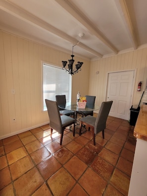 A dining area with a glass table, four chairs, a chandelier, a window with blinds, and a door with a fire extinguisher. - Vero Beach Hideaway- 5-minute drive from beach (Vero Beach)