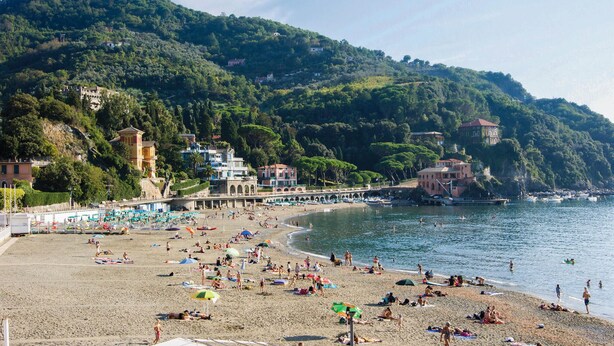 Une plage où l’on peut voir des gens se prélasser au soleil et nager, une jetée avec des bâtiments, et des collines en arrière-plan.