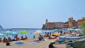 Una playa con gente tomando el sol y nadando, sombrillas coloridas y barcos en el agua.