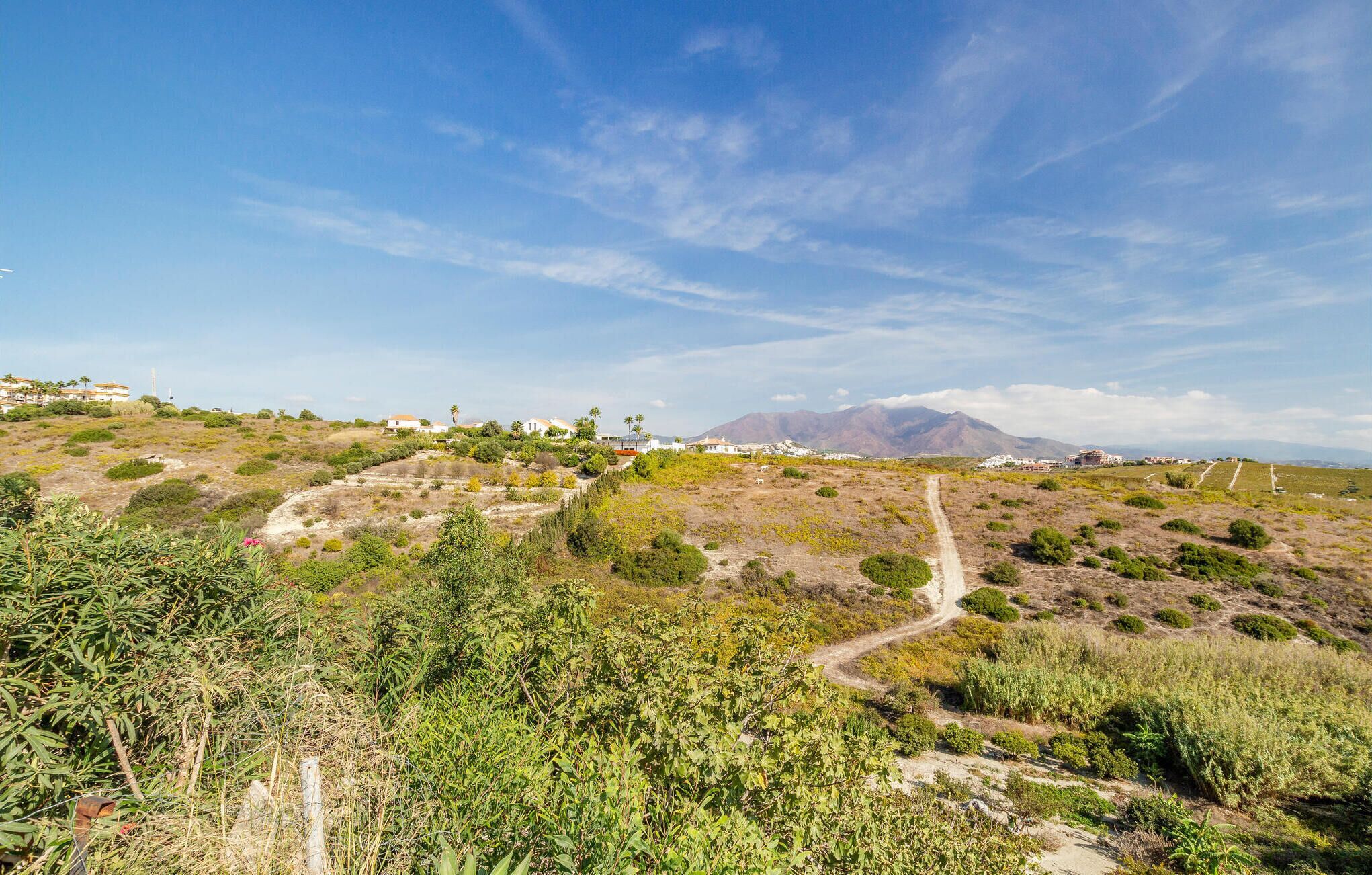 A hilly landscape with a dirt path, vegetation, and distant buildings.