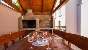 Un espace de restauration en plein air doté d’une cheminée en pierre, de bancs en bois et d’une table dressée pour quatre personnes, avec des verres à vin.