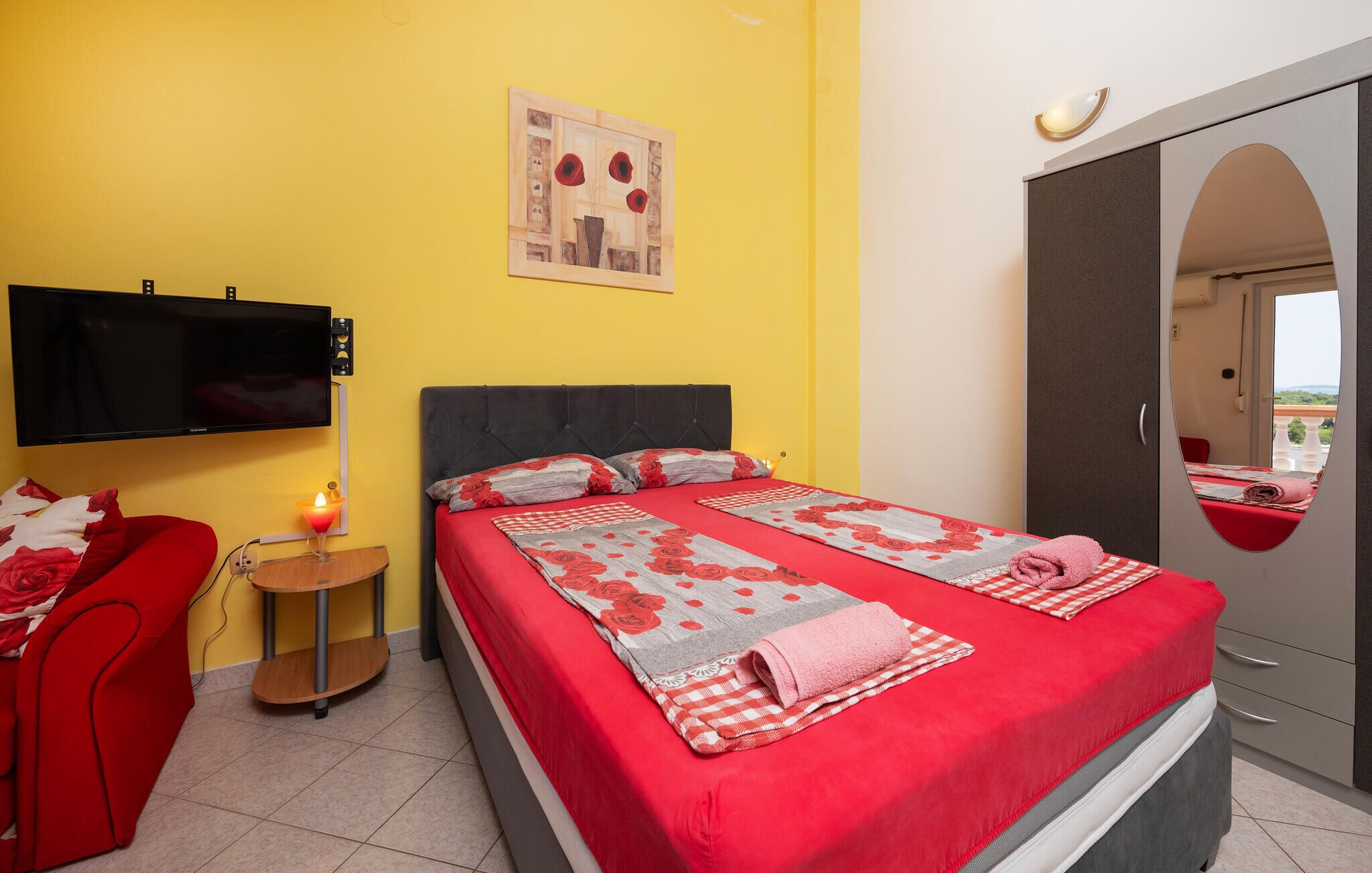 Une chambre d’hôtel avec un lit, une télévision, une petite table avec une bougie, une chaise rouge et une armoire avec un miroir.