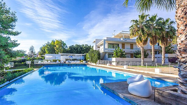 Une piscine dotée d’une terrasse en bois et de chaises longues, entourée de palmiers et bordée d’un bâtiment moderne avec des balcons.