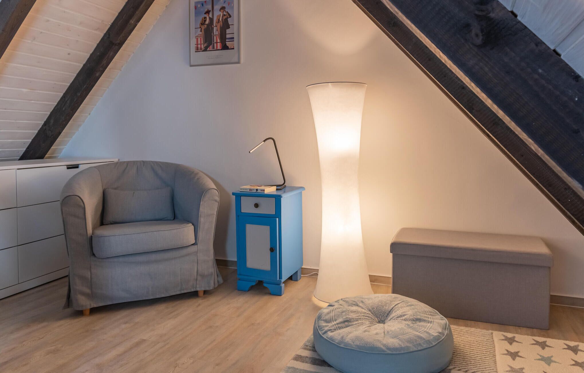 A cozy loft bedroom with a sloped ceiling, a grey sofa, a blue nightstand, a tall floor lamp, and a small ottoman.