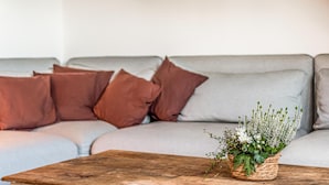 Een woonkamer met een grijze bank, bruine kussens en een houten salontafel met een gevlochten mand vol witte bloemen.